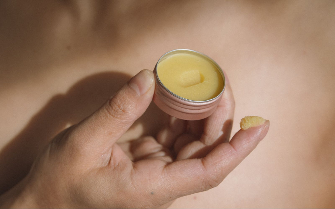 An open 15g tin of Universal Balm held in one hand, with a small scoop of balm on a fingertip